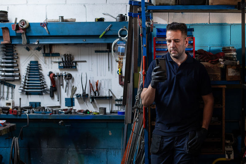 Serious adult male mechanic in workwear standing near shelves with professional tools and instruments and checking information on smartphone while working in workshop