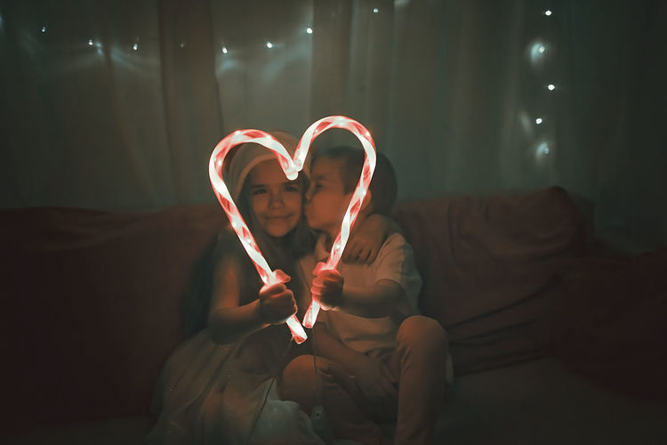 Siblings making heart shape of illuminated candy canes on sofa in darkroom