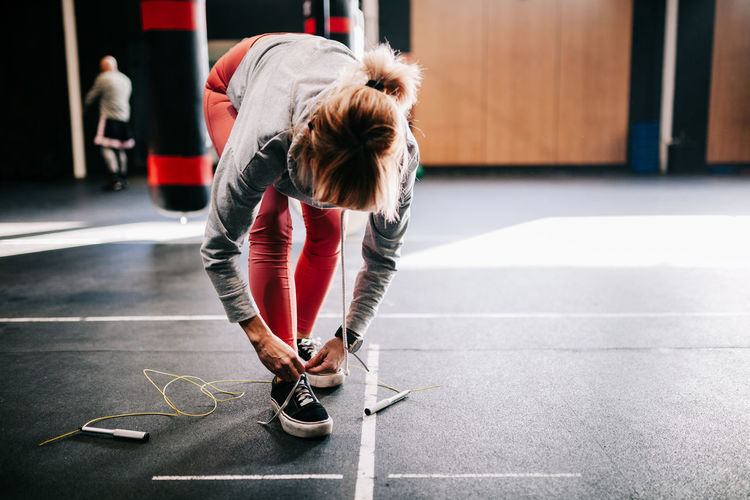 Unrecognizable female athlete in sportswear tying laces on sneakers while standing near hanging punching bag and preparing for workout in modern gym