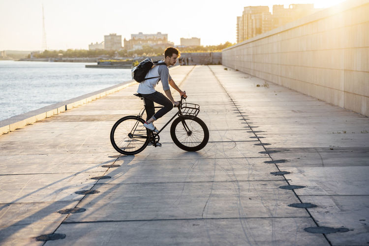 Young man with backpack riding bike on waterfront promenade at the riverside at sunset