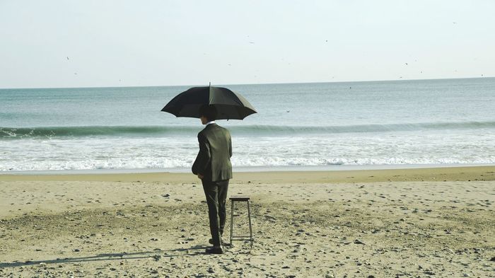 Rear view of well-dressed man holding umbrella at beach against sky