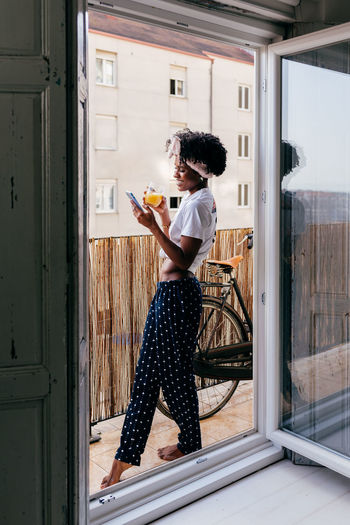 Optimistic african american lady with glass of juice browsing smartphone on balcony in morning