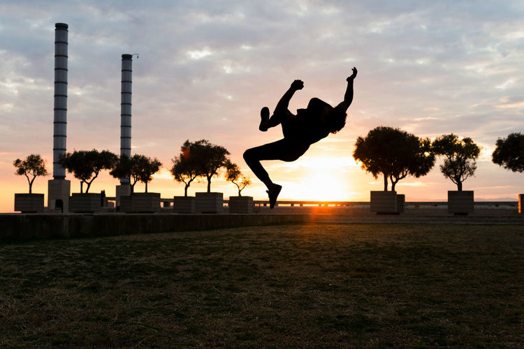 Full body of unrecognizable male silhouette jumping above ground with raised arms while doing parkour trick on sundown