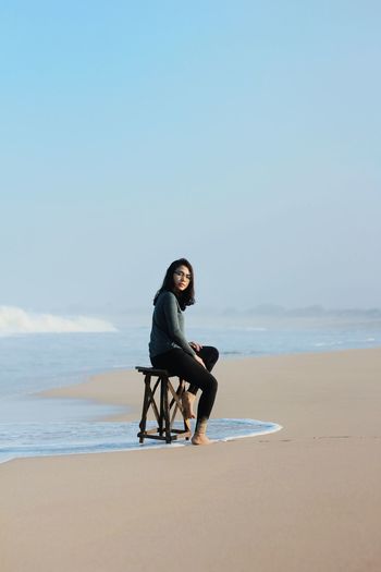Side view of woman sitting on seat at beach against blue sky