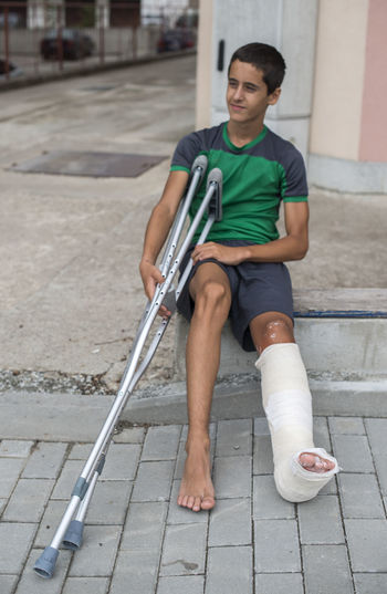 Boy with broken leg holding walking cane while sitting on footpath