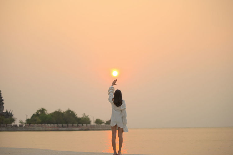 Rear view of woman standing on shore against sky during sunset