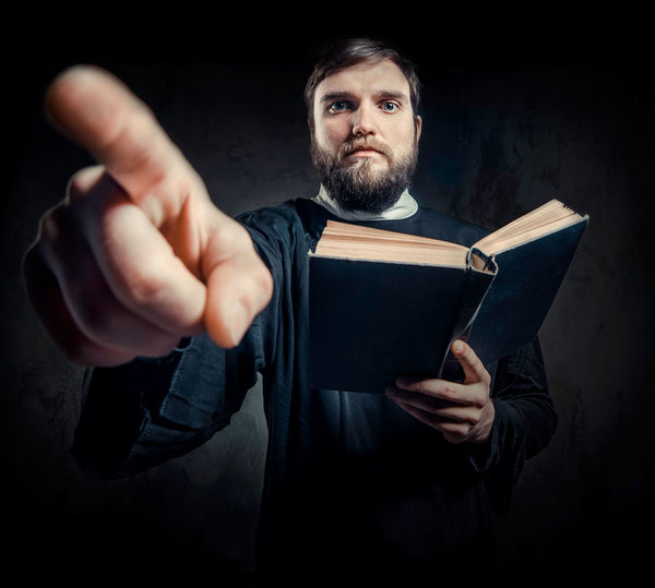 Portrait of priest pointing while holding bible against black background