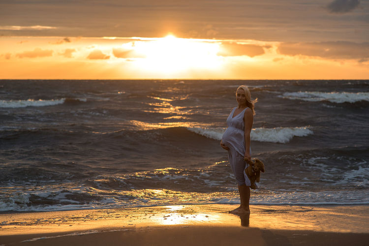 Pregnant woman standing on beach against sky during sunset
