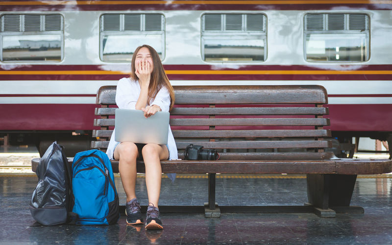 Happy woman using laptop while sitting on bench at railroad station platform