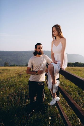 Bearded man standing by woman sitting on railing against clear sky