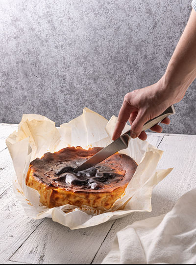 Woman is cutting homemade basque burnt cheesecake in baking paper on textured white wooden table. 