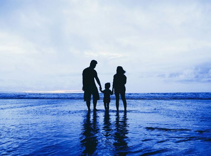 Silhouette family standing on shore at beach against cloudy sky