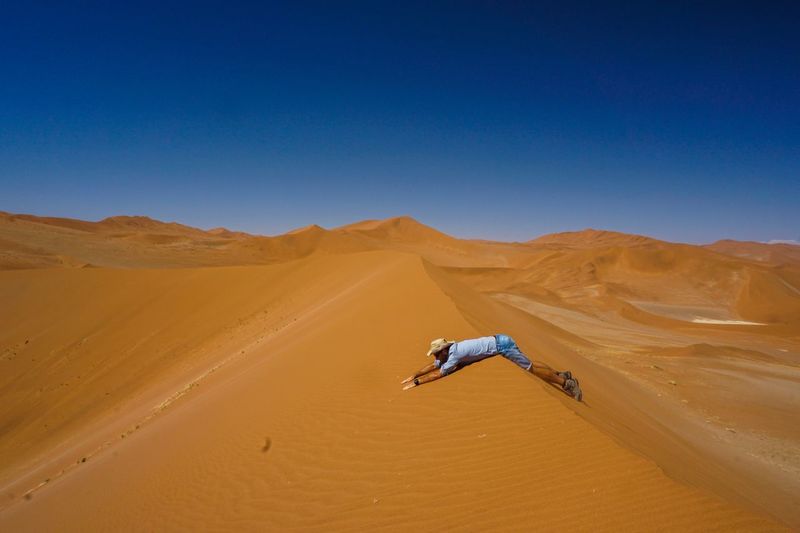Side view of man lying on sand dune in desert against clear blue sky