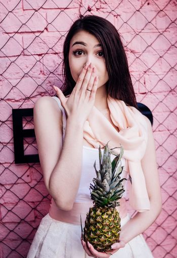 Portrait of beautiful woman holding pineapple while standing against pink wall