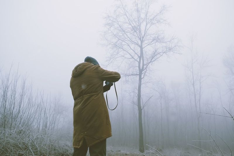Rear view of man standing against bare trees in foggy weather