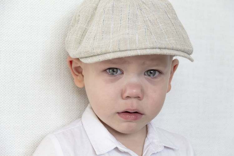 A small, sad boy in caps with huge tears in his eyes stands near a white wall.