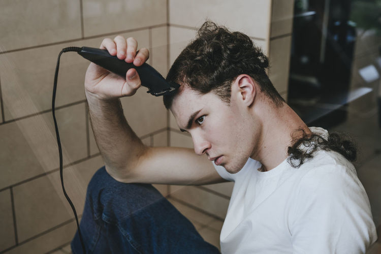 Sad man cutting his own hair with electric razor at home