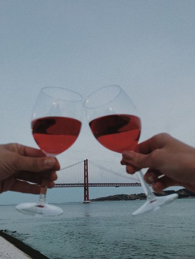 Cropped hands toasting wineglasses over sea against clear sky during sunset