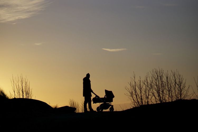 Silhouette man with baby stroller standing against sky during sunset