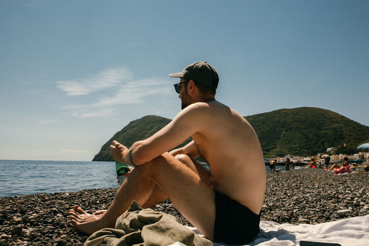Full length of shirtless man sitting on beach against sky