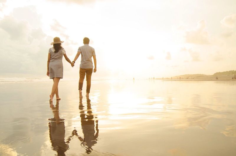 Rear view of couple walking at beach against cloudy sky during sunset