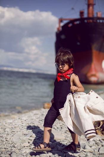 Fashionable boy child with long hair stand on a log next to a large ship that ran aground 