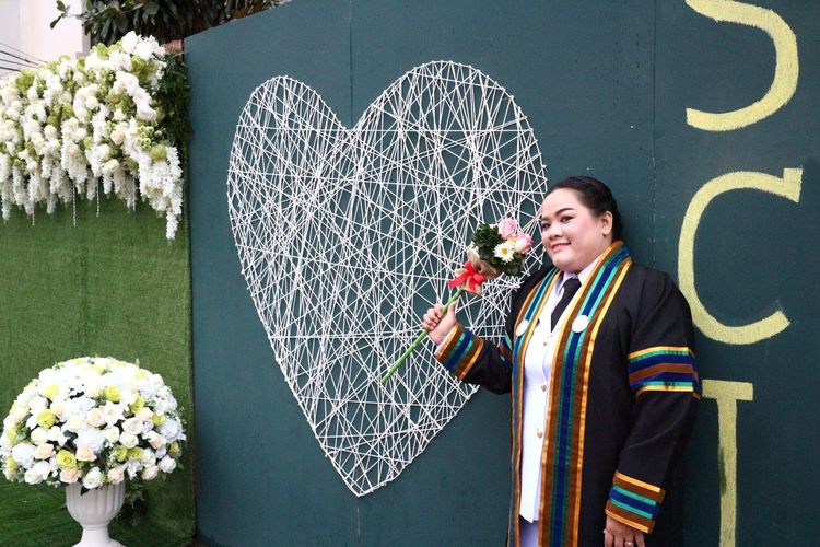 Portrait of overweight smiling woman wearing graduation gown while standing by heart shape