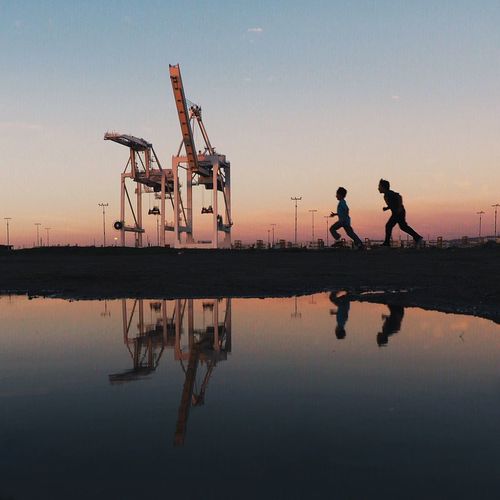 Reflection of boys running by calm lake against crane during sunset