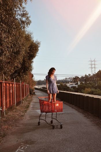 Young woman standing in shopping cart on footpath against sky