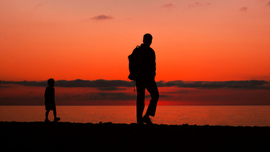 Silhouette men walking on beach against orange sky