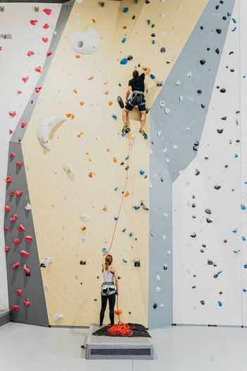 Back view of anonymous woman in activewear helping anonymous male alpinist to climb artificial wall during training in modern gym