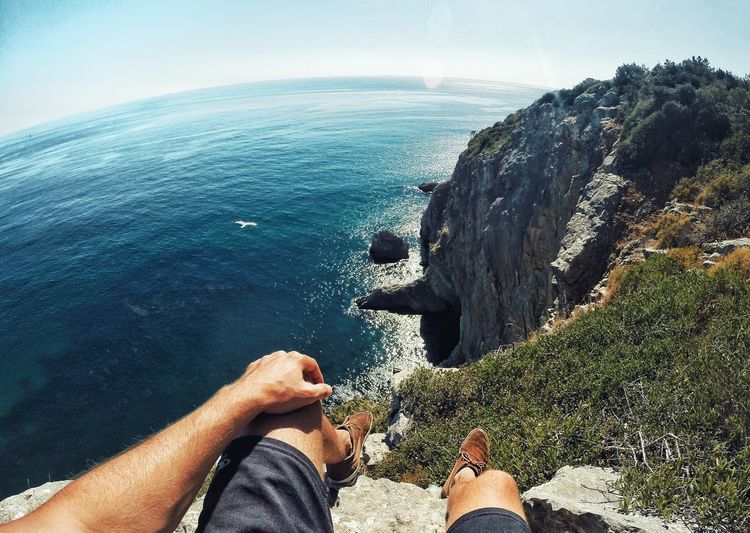 Low section of man sitting on cliff by sea against sky