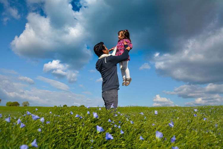 Father carrying daughter by blooming flowers against sky