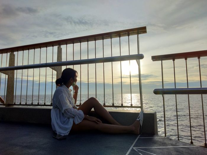 Side view of woman sitting on railing against sky