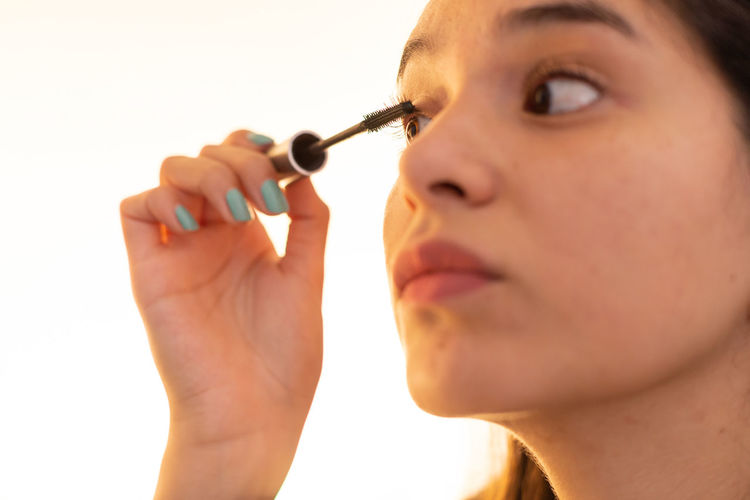 Young ethnic lady looking in mirror while applying mascara