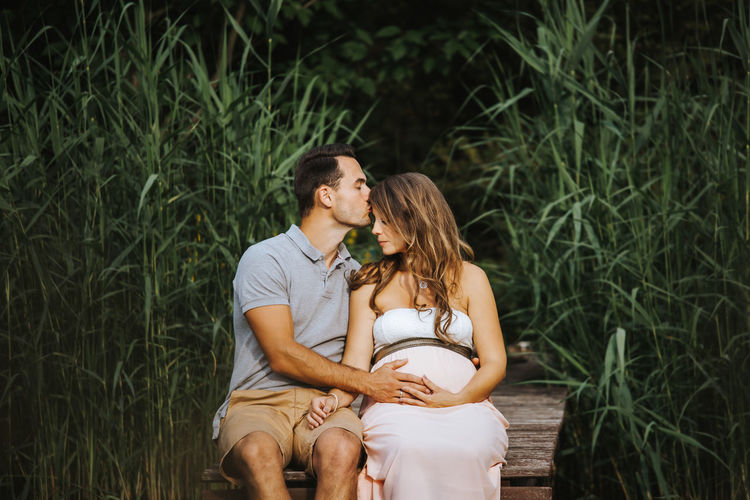 Man kissing pregnant wife while sitting on pier over lake
