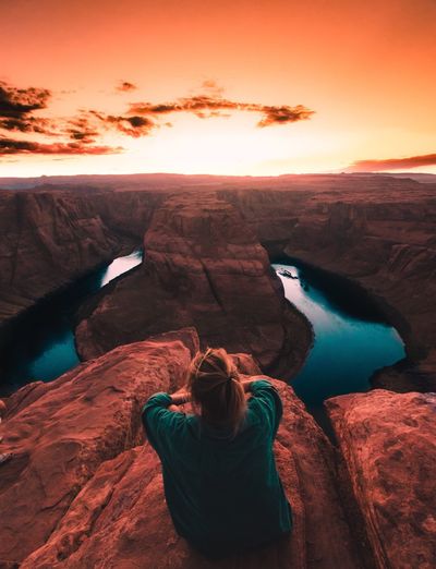 Rear view of woman sitting at horseshoe bend against orange sky