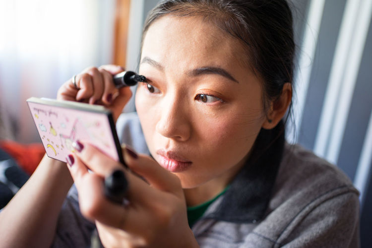Young asian female in casual clothes with small mirror in hand doing makeup and putting black mascara on eyelashes