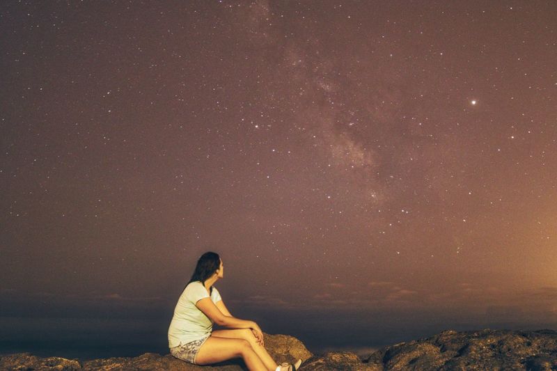 Side view of woman sitting on rock against sky at night