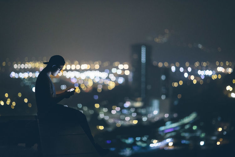 Side view of man using phone while sitting on retaining wall in city at night