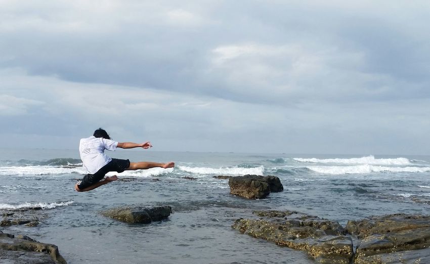Rear view of mid adult man jumping by sea against cloudy sky
