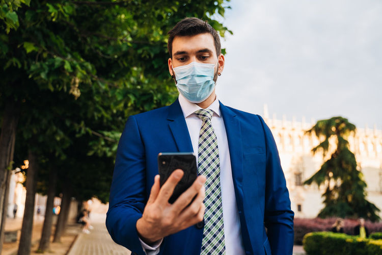Young elegant businessman with surgical mask looks at phone in park