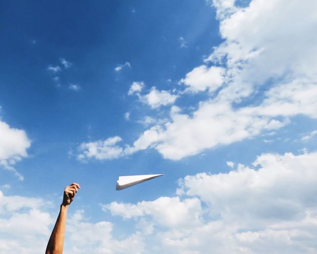 Cropped image of hand flying paper airplane against sky
