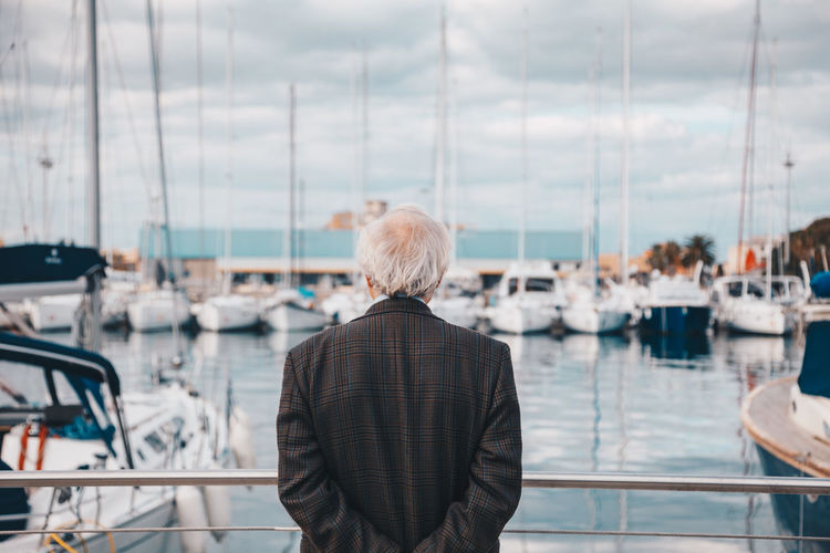 Rear view of senior man with hands behind back standing by railing at harbor