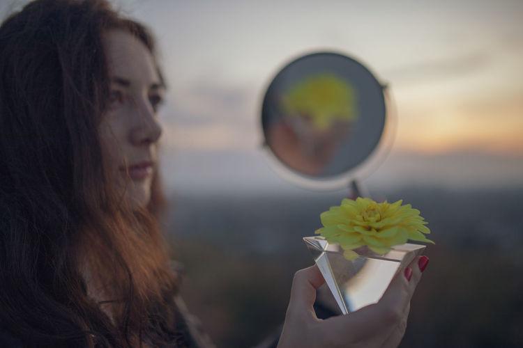 Woman holding hand mirror with reflection of crystal and flower during sunset