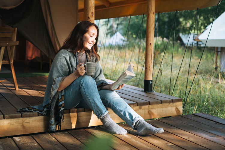 Young brunette woman in poncho reading book and relaxing in glamping in the woods