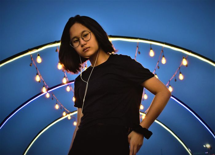 Young woman looking away while standing against illuminated lights