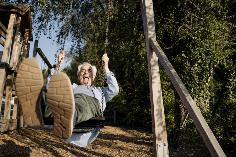 Senior man sticking out tongue while playing on swing at park