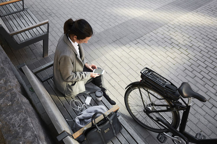 High angle view of woman surfing net through smart phone while sitting on bench in city