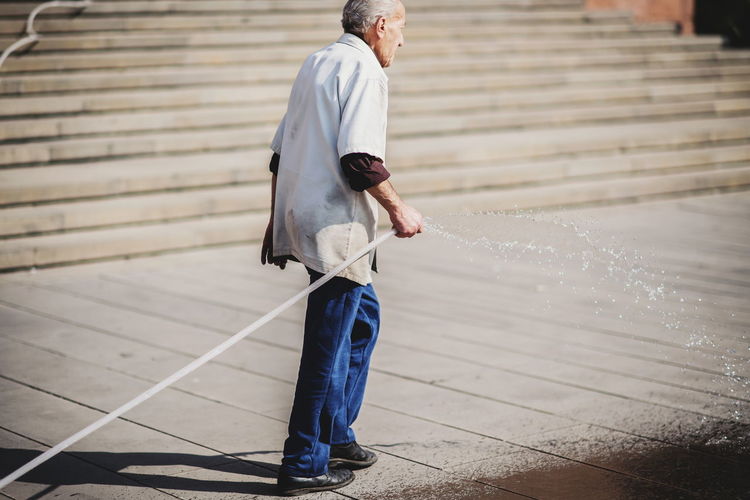 Full length of senior man cleaning street using garden hose in city